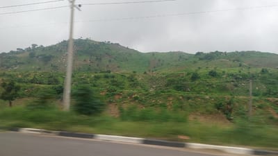 The image captures a vast, rolling green hillside under an overcast sky, viewed from a roadside perspective. The hillside is dotted with rocks and sparse vegetation, with a more densely vegetated area in the foreground. A utility pole, likely supporting power lines, stands prominently on the left side of the frame, near the center. The foreground shows a blurred asphalt road with a distinctive black and white striped curb, indicating motion and suggesting the image was taken from a moving vehicle. The scene appears to be located in Dam Camp, Nigeria, a region known for its hilly terrain.