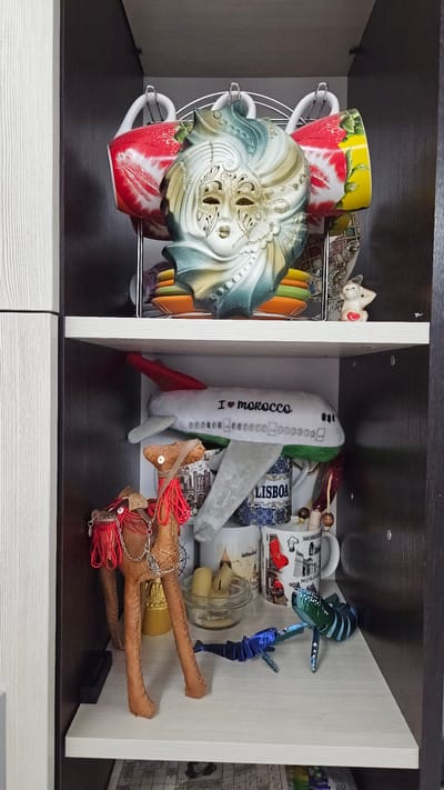 The image shows the interior of a dark brown and white shelf or cabinet. The shelf is divided into two compartments. The left compartment contains a metal mug rack with several colorful mugs, a decorative mask and a small ceramic figurine. The right compartment contains a plush airplane with "I heart morocco" written on it, several mugs with various city names printed on them, a jar with objects inside, a blue and green fish ornament, and a leather camel figurine with red tassels. The environment is indoor and appears to be a storage space. No people are visible. The location of the scene is presumed to be in Yerzovka, Russia.