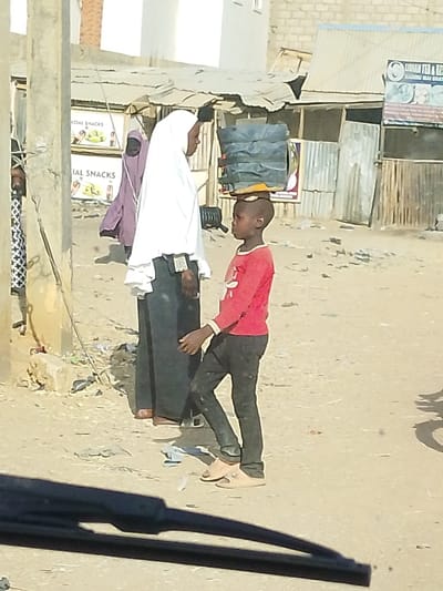 A boy with black pants and a red shirt carries a stack of containers on his head. He wears light-colored sandals and stands next to a woman in a white hijab and dark skirt. They are on a dirt road in front of buildings, some with signs that say "Snacks" and another with "Tea & Snacks." A second woman in a purple robe stands in the background, with trash visible on the ground. A car windshield is visible at the bottom of the frame.