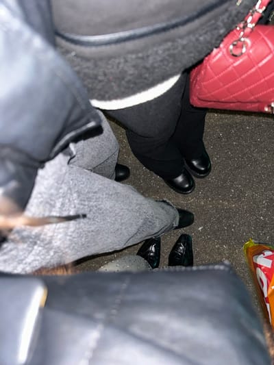 A top-down shot reveals three pairs of legs and shoes on a dark surface. The left features black pants and chunky black shoes, alongside a red quilted purse. Adjacent, the second pair wears grey jeans and black shoes. To the right, the final pair sports black, shiny shoes and a lighter colored ankle with some ornamentation. A large black bag dominates the right side of the frame. An orange and white snack bag sits in the upper right corner.