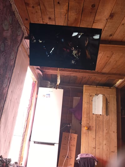 The image depicts the interior of a room with a wooden plank ceiling and walls. A flat-screen television is mounted on the ceiling, displaying a dark scene with illuminated elements. To the left, a large window is partially covered by a patterned curtain. In the center of the room stands a white refrigerator, and to its right is a rustic wooden door with a small arched mirror embedded in it. A brown cardboard box and some clothing items are visible on the floor. The overall impression is of a simple, lived-in space, possibly a rural dwelling or a room in a traditional wooden building. The location is identified as Monastirische, Russia.