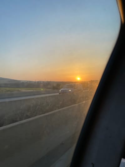 This is a photograph taken from inside a vehicle, looking out at a highway at sunset. The sky is a gradient of blue to orange, with the sun low on the horizon. There are cars on the highway, and in the distance, there are buildings and hills. The scene is likely in or near Ouled Djilali, Algeria, given the context. The foreground is dominated by the interior of the vehicle, with a dark, curved edge on the right side of the frame, likely a car window frame. The middle ground shows a concrete divider on the highway, and beyond that, the road with vehicles. The background features a rural landscape with some development. The overall impression is one of travel at dusk, with the warm glow of the setting sun illuminating the scene.