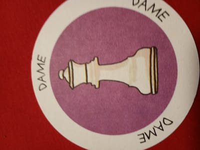 The image shows a circular playing card with a drawing of a queen chess piece in the center, set against a purple background. The word "DAME" is written in black text around the perimeter of the card, with the letters oriented to be read in different directions. The card itself is white with a black outline. The card is placed on a red surface, which appears to be a table or a tablecloth. There is no indication of the city of Rosenheim, Germany, in the image.