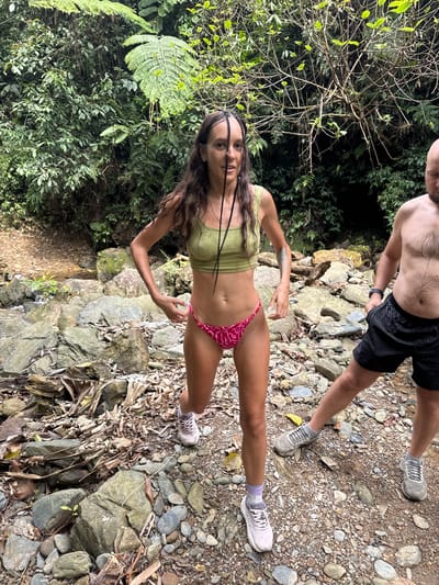 A woman and part of a man are in a rocky, tropical area. The woman stands facing the camera, wearing a light green top and a pink patterned bikini bottom. She wears sneakers and has braids in her hair. The man, visible from the waist up, is shirtless and wears black shorts. The ground is covered with rocks, leaves, and foliage. Green tropical plants and trees surround the area. The image is taken outdoors. The specific location in Suối Giàng, Vietnam, is not discernible from the image.
