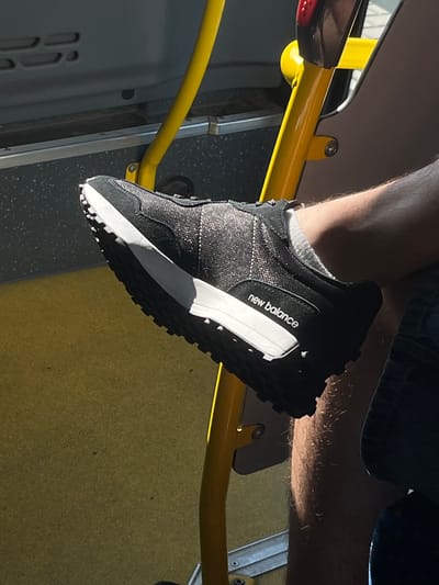 The image shows a close-up view of a person's foot wearing a black New Balance sneaker. The shoe has a speckled, shimmering upper and a thick white sole with black accents. The sneaker is resting on a yellow metal pole, possibly a handrail on public transportation. The background includes a yellow surface that appears to be a seat or floor and a dark gray panel. Sunlight is casting shadows, suggesting it is daytime. The scene appears to be indoors, possibly on a bus or train, given the presence of the handrail and the utilitarian interior. The overall impression is casual and candid. The text "new balance" is clearly visible on the side of the shoe. The location context is Dobřany, Czechia.
