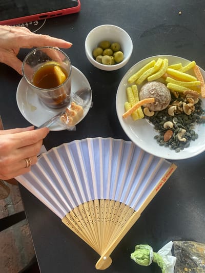 The image shows a close-up, overhead view of a table with a selection of snacks and a drink, with a hand reaching for the drink. In the foreground, a white and light blue foldable fan is laid out. A red smartphone case is visible in the upper left corner of the image. In the center of the image, a glass filled with a dark liquid and a slice of citrus fruit sits on a white saucer. A spoon and a small, individually wrapped pastry are on the saucer next to the glass. To the right of the glass, a white plate contains a variety of snacks, including olives in a small white bowl, various nuts and seeds, what appears to be a biscuit or cookie, and some long, thin, yellow and orange crackers. The table surface is black. The fan has "Adrián, M. Alcalde" printed in red on one of its wooden ribs, indicating a location in San Pedro de Alcántara, Spain.