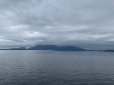The image shows a calm body of water in front of a mountainous landscape under a cloudy sky. The water is dark and reflects the muted tones of the sky. Gentle ripples can be seen on the surface. In the background, a range of mountains stretches across the horizon, with the peaks of some of the closer mountains partially obscured by low-hanging clouds. The overall impression is one of a serene, slightly somber, and vast natural setting. The light suggests it is daytime, but the heavy cloud cover creates a diffused and subdued atmosphere. The landscape appears to be in a coastal or fjord region, characteristic of the Sandnessjøen, Norway location. There are no visible people or man-made objects, emphasizing the natural beauty of the scene. The weather is overcast, contributing to a peaceful yet potentially melancholic mood.