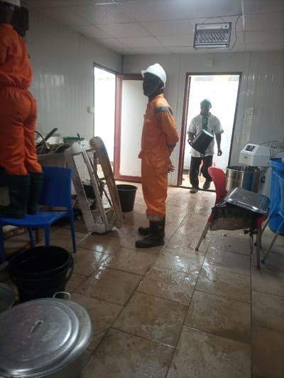 The scene depicts an interior space, likely a commercial kitchen or utility room, within the city of Opete, Nigeria. The tiled floor is visibly wet across the entire foreground and midground. Three adult males are present.
On the left, partially visible, a person wearing an orange coverall and dark rubber boots stands on a blue plastic chair. In the center, a male with dark skin wears an orange coverall with grey shoulder accents, a white hard hat, and dark rubber boots. He stands upright, facing right, with his hands clasped behind his back, observing the environment. Through an open doorway on the right, another male with dark skin is entering the room. He wears a white short-sleeved top with a patterned design, dark trousers, and a light blue head covering. He carries a black bucket, tilted forward, in his left hand.
Various objects are distributed throughout the wet room. A large white industrial fan lies on its side on the floor near the center. Several black plastic buckets are visible; one, filled with liquid, is on the floor near the blue chair, and another is near the feet of the central figure. In the foreground, left, several large silver-colored metal pots with lids are on the floor. On the right, a red plastic chair holds a silver metal food tray. Behind it, a white industrial kitchen mixer with a large bowl attachment is visible on a surface. A black electronic insect zapper is suspended from the white ceiling. The walls are constructed from white modular panels.