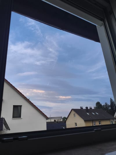 A view through a window in Pegnitz, Germany, shows a partly cloudy sky with a thin contrail from an airplane. The sky is a mix of blue and gray, with some lighter pinkish hues near the horizon, suggesting it is either early morning or late evening. Below the sky, the rooftops of several houses are visible. On the left, a white house with a red-tiled roof has a single dark-framed window. To the right of this house, a yellow house with a dark-tiled roof has multiple windows and two chimneys. The scene appears peaceful and residential. The time of day is likely dusk or dawn due to the lighting and sky conditions. No people or text are visible in the image.