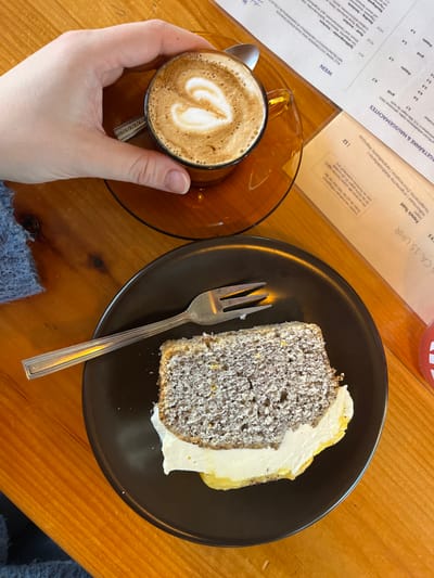 An overhead photograph depicts a tabletop scene inside a cafe or restaurant located in Kaiserslautern, Germany. In the upper left quadrant, a human hand, presumed female, holds an amber-tinted glass cup containing a latte with heart-shaped art on its foam. The cup rests on a matching saucer, which also holds a small silver spoon. A portion of a blue knitted garment is visible on the left, belonging to the individual whose hand is shown.
In the lower right quadrant, a slice of cake is presented on a dark brown, matte ceramic plate. The cake is light greyish-brown with visible yellow flecks, topped with a thick layer of white frosting and a yellow curd or cream layer beneath it. A bite has been removed from the cake's left side. A silver-colored metal fork rests on the plate to the left of the cake.
The table surface is made of light-to-medium brown wood with visible grain. To the right of the food items, a laminated menu in German is visible, showing sections like "WEIN" and "GETRÄNKE & HAUSGEMACHTES" with prices in Euros. Behind this menu, a white sheet of paper confirms the location with "KAISERSLAUTERN" printed at the top, along with other German text including "French Toast" and "CA. 15 UHR". A small, indistinct red object is partially visible on the far right edge of the table.