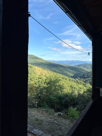 The image shows a view of rolling, forested hills under a bright blue sky with scattered white clouds, as seen from the interior of a dark, enclosed space. The foreground is dominated by a dark, wooden structure, likely a porch or balcony, with a thick, dark post on the left and a sloping wooden beam across the bottom right. A thin wire stretches diagonally across the upper portion of the frame, with a black clothespin attached to it. The background reveals a vast landscape of green hills layered with varying shades of green foliage, indicating a dense forest. The sunlight casts shadows across the slopes, highlighting the undulating terrain. In the distance, successive layers of mountains fade into a hazy blue, suggesting a wide expanse of undeveloped natural land. The location is identified as Manaselska Reka, Bulgaria, a region known for its mountainous and forested terrain. There are no people or animals visible in the image. The scene depicts a tranquil, natural landscape viewed from a sheltered vantage point.