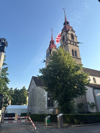 The image captures a clear, sunny day in Winterthur, Switzerland, with the sky a vivid blue. The scene is dominated by the towering spires of a historic church, its stone facade and red-tiled roofs prominent against the sky. Two flags, one bearing a red heraldic symbol on a white background, are visible fluttering from the church towers. Lush green trees partially obscure the building, adding a touch of nature to the urban landscape.
In the foreground, a large construction crane is partially visible, suggesting some form of work or maintenance is underway. To the right, a cobblestone street is lined with hedges and a metal bin. Red and white striped barriers, marked with the word "STUTZ," indicate a temporary closure or cordoned-off area. A white tent structure is also visible, further hinting at an event or ongoing construction. The overall atmosphere is one of a bright, active day in a picturesque Swiss city.