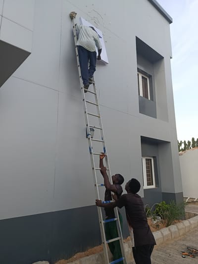 Two men are ascending a metal ladder attached to a modern, grey building. One man, higher up on the ladder, is wearing a light-colored shirt and dark pants and is reaching to attach a white rectangular sign to the building's facade. Below him, a second man in a dark shirt and pants is also on the ladder, holding it steady. A third man, also in a dark shirt and pants, is standing on the ground at the base of the ladder, looking up. The building features dark gray trim and windows with white frames. The ground is covered in a mixture of sand and paving stones, with some vegetation visible in the background. The sky is overcast. The scene is likely in Dutse, Nigeria, given the architecture and the presence of workers installing signage, a common activity in developing urban areas.