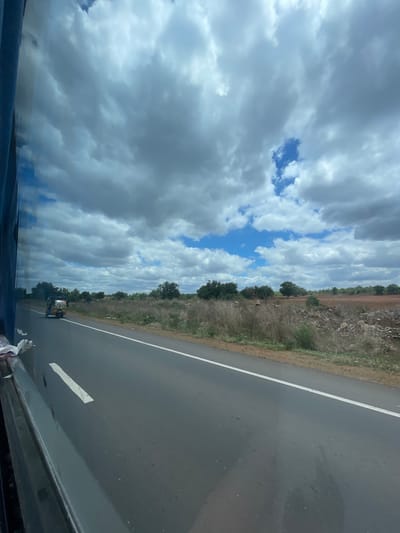 A view from inside a vehicle looking out at a two-lane road in Sugali Tanda, India. The road is paved and has a white line marking the lane. A yellow and blue autorickshaw is traveling on the road in the distance. The landscape beside the road is dry with sparse vegetation and scattered rocks. In the background, there are trees and a red, rocky field. The sky is filled with dramatic, gray and white clouds, with patches of blue sky visible. The reflection of the vehicle's interior is visible on the left side of the image.