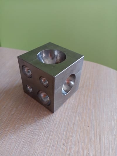 A metallic cube with indentations is placed on a light-colored wooden surface. The cube has a hemispherical indentation on its top face and circular indentations on its side faces. The background is a plain light green wall. The scene is indoors. There are no people or animals present. The image is a close-up shot of the object. The location is Vilyuysk, Russia.