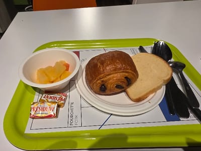 A green tray holds a breakfast meal. A white bowl contains cut fruit, alongside two small packets of President butter. A white plate holds a pain au chocolat and a slice of white bread. To the side are a knife, fork, and spoon. The tray sits on a white surface.