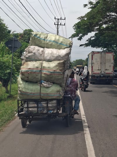 The image shows a motorcycle pulling a trailer loaded with large sacks, presumably in Keudealuerheung, Indonesia. The motorcycle driver, wearing a red and white shirt and blue jeans, appears to be lying on the pile of sacks. The trailer is constructed of a metal frame and contains more sacks inside. Behind the motorcycle are two other motorcycles and a truck. The road is paved with a white line. Trees are visible on either side of the road, and there are power lines above.