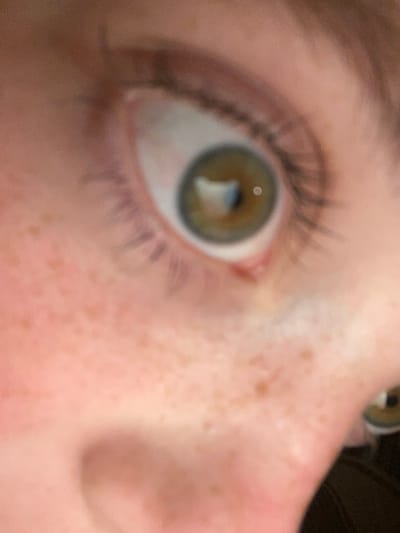 This is a close-up, macro photograph of a person's eye. The eye is the central subject, with the iris appearing to be a hazel or green-brown color. The sclera, or white of the eye, is visible, as are the eyelashes. The skin surrounding the eye is fair and exhibits a scattering of freckles. The focus is sharp on the eye, with the rest of the image softly blurred, suggesting a shallow depth of field. The lighting appears to be natural, possibly from an indoor source or during daylight hours. There are no visible text elements in the image. The framing and extreme close-up suggest an intentional focus on the details of the eye and surrounding skin.