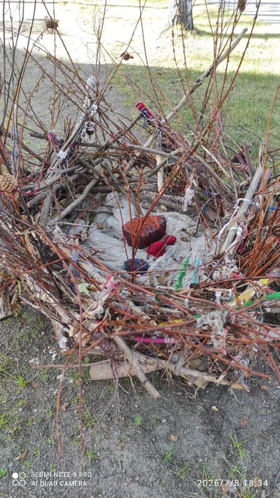 The image displays an outdoor scene in Iisalmi, Finland, featuring a large, rustic structure composed of numerous branches and twigs. The structure appears to be woven or piled together, forming a somewhat hollow interior. Inside this structure, there are several balls of yarn in various colors, including orange, red, and blue. A piece of light-colored fabric is visible beneath the yarn. The exterior of the structure is decorated with various colorful scraps of fabric and ribbons tied to the branches. Some pinecones are also incorporated into the arrangement. The ground surrounding the structure is covered with gravel, dirt, and sparse green grass. In the background, more grass and the trunk of a tree are visible. The lighting suggests it is daytime.