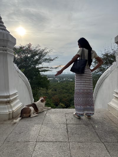 A woman with long dark hair, wearing a light-colored t-shirt and a long patterned skirt, stands with her back to the camera, facing a cityscape and lush green landscape. Her left arm is extended outwards, as if gesturing or reaching. She carries a dark tote bag over her left shoulder. To her left and slightly in the foreground, a brown and white dog lies on a stone surface, facing away from the woman and looking towards the landscape. The scene is set on a tiered stone structure with ornate white architectural elements on either side. In the background, the sun is partially obscured by clouds, casting a soft light on the scene. Distant buildings are visible on the horizon, nestled amongst a dense expanse of trees. The overall impression is one of a tranquil moment overlooking a city and natural surroundings, possibly at an elevated viewpoint such as a temple or observation point within the มาบฟักทอง, Thailand area.
