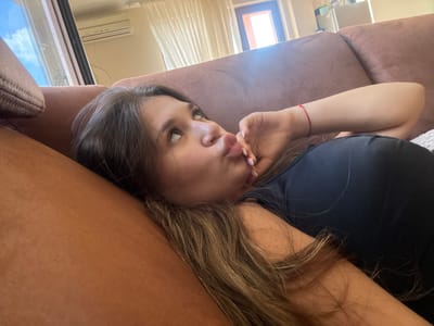 This media file depicts a young woman lying on a couch, with her head resting on a pillow. She is looking upwards and to her right, with her lips pursed in a playful expression. Her right hand is raised to her face, touching her cheek. She is wearing a black top, and a thin red string bracelet is visible on her wrist. The couch appears to be made of a textured, light brown material. In the background, a window is visible with white curtains, and sunlight streams through, suggesting it is daytime. An air conditioning unit is mounted on the wall above the window. Further in the background, what looks like kitchen cabinets and a refrigerator can be seen, indicating a domestic setting. The overall scene conveys a relaxed and casual atmosphere. No visible text is present in the media file.