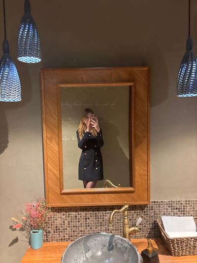 This image depicts a female subject taking a mirror selfie in a restroom or washroom facility. The location is identified as Hinkalnaya No.1 Restaurant, situated at Ulitsa Voroshilova, 137, Serpukhov, Russia.
The primary subject, reflected in a large rectangular mirror with a thick light-wood frame, is a woman with long, light-brown hair. She is wearing a dark, double-breasted dress featuring two vertical rows of gold-colored buttons, along with dark tights. Her hands are raised, holding a light-colored smartphone to capture the reflection.
Above the mirror, two light blue, woven-textured pendant lamps hang from the ceiling, illuminated from within. The walls are a uniform, plain olive-green or dark beige color. Directly behind the woman's reflection, on the wall inside the mirror's view, several lines of cursive Georgian script are visible, including the phrase "გემრიელი ბროწეულის წვენები" (Delicious pomegranate juices).
Below the mirror, a wooden countertop supports a dark, irregular-shaped stone basin. To the right of the basin, an antique-style brass faucet with a white handle accent is installed. Further to the right on the countertop, a dark, rectangular soap dispenser with a brass pump is positioned next to a woven brown basket containing folded white paper towels. To the left of the basin, a small teal vase holds a collection of dried flowers and twigs, predominantly in shades of pink and muted brown. The wall immediately beneath the countertop is covered in small, square mosaic tiles in various shades of brown and beige.