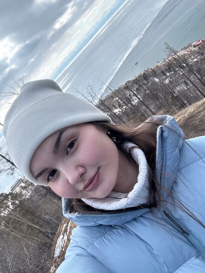 A selfie-style image captures a young female in the foreground, positioned at an elevated vantage point. She has fair skin, dark eyes, and dark brown hair, which is visible beneath a light gray knitted beanie. She wears a light blue puffer jacket over a white, possibly knit, high-collared garment. Her expression is neutral with a slight smile.
The background reveals a panoramic view of a large body of water, likely Lake Baikal, extending to the horizon under a partly cloudy sky. The water exhibits a calm surface, reflecting light, with potential ice formations or bright reflections. A small, dark-colored boat is visible on the water in the mid-distance. The shoreline on the right features a wooded area with numerous bare, leafless trees, and patches of snow are visible on the ground, indicating a cold weather season. Several buildings with reddish roofs are discernible amidst the trees along the distant shore. The overall scene suggests a location in Nikola, Russia, overlooking Lake Baikal.