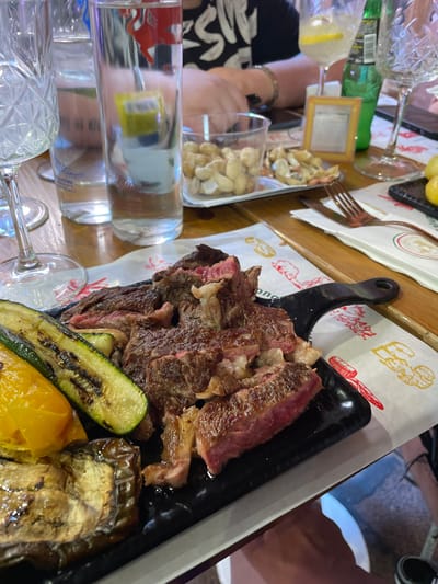 A high-angle shot depicts a dining table setting. A black rectangular serving platter takes central focus, laden with sliced steak, grilled zucchini, grilled eggplant, and yellow peppers. The meat displays a gradient of cooked hues from pink to brown. The platter rests on a white paper liner imprinted with cartoon-style pig and meat product illustrations. Behind the platter, a fork lies beside a small dish of white food items. Transparent glass bottles and drinking glasses are placed in the foreground, refracting light. Visible on the left are people's arms and clothing, indicating diners. The background exhibits a mix of objects including more dishes. Bologna, Italy is not specifically confirmed in the image.