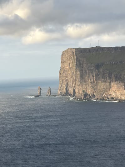 The image depicts a dramatic coastal landscape with a towering cliff face dominating the right side of the frame. The cliff is a sheer, rugged wall of rock, showing distinct layers and textures, with patches of green vegetation clinging to its upper edges. Two tall sea stacks, also rock formations, rise from the dark blue ocean in the middle ground, standing as solitary sentinels against the vast expanse of water. The sea is choppy, with whitecaps visible near the shore and around the base of the sea stacks. The sky above is overcast, with heavy gray clouds interspersed with brighter, illuminated patches, suggesting a diffused light source. Two slender, tall antenna structures are visible on the plateau at the top of the cliff. The overall scene conveys a sense of wild, untamed nature. The location is the coastline near Tjørnuvík, Faroe Islands, a region known for its dramatic cliffs and sea stacks.