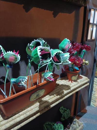 An indoor scene in El Port De Sagunt, Spain, displays a collection of art made from recycled materials. The primary focus is a brown rectangular planter containing several stylized "carnivorous plants," constructed from cut green and transparent plastic bottles. These plants feature jagged white "teeth" and pink interiors, mounted on white and green stems. To the right, a smaller orange pot holds red, rose-like flowers, also appearing to be made from cut plastic. Both containers rest on a shelf composed of horizontally arranged wine corks. The background consists of a plain brown wall. To the far right, a miniature building facade is visible, featuring a brown-framed window with white panes and a brass-colored doorknob-like fixture positioned below the red flowers. Below the cork shelf, on a light blue-grey surface, are green, leaf-like sculptural elements. No people are present.