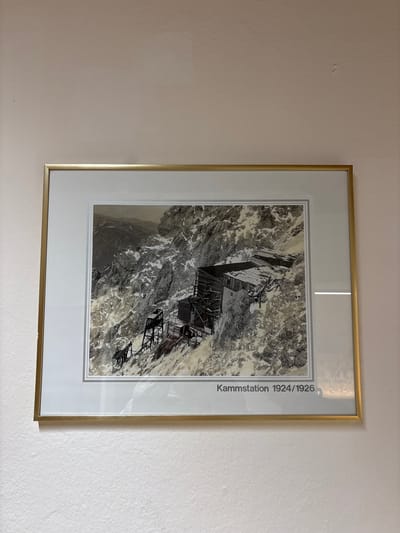 The image shows a framed black and white photograph hanging on a wall. The photograph depicts a mountainous scene with snow-covered slopes and several wooden structures, possibly buildings or stations, built on the mountainside. There are also cables and supports visible, suggesting some form of transportation system like a cable car or railway. The text below the photograph reads "Kammstation 1924/1926". This likely refers to the "Kammstation", a mountain station, with the dates indicating when the photograph was taken or the structures were in place. The location is identified as Ehrwald, Austria, which is a town known for its mountainous terrain and ski resorts. The overall scene is one of a historical record of infrastructure development in a mountainous region.