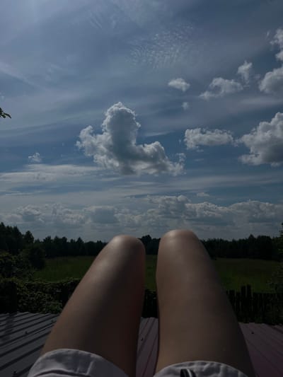 The image is a first-person perspective shot looking upwards at the sky, with a person's legs visible in the foreground. The legs are bare and appear to be positioned on a dark, corrugated surface, likely a roof. Beyond the legs, a band of dark green trees forms a horizon line, with a field of green grass and some indistinct structures behind it. The sky is a prominent feature, filled with a mix of white, fluffy clouds and thinner, wispy clouds against a blue sky. The lighting suggests it is daytime, with sunlight illuminating the scene. The overall impression is one of relaxation and observation of the sky. The location in the city of Kemul, Russia, is not explicitly visible within the image itself, but the description is given as context.