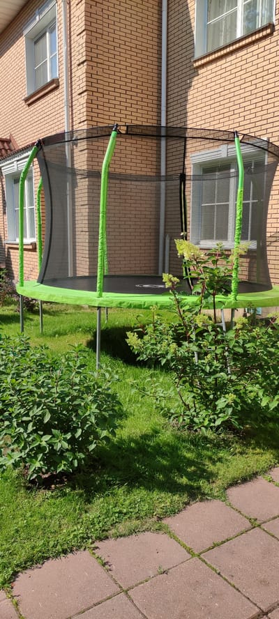 In the city of Rosinka, Russia, a large, circular trampoline with green padding and a black safety net is positioned on a grassy lawn. The trampoline is situated in front of a brick building with multiple windows. Lush green bushes flank the trampoline and a paved patio area made of reddish-brown tiles is visible in the foreground. The sun is shining, casting shadows across the scene.