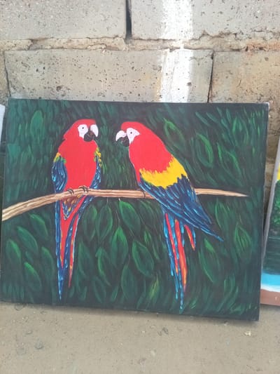 A painting of two red and blue macaws perched on a branch is leaning against a cinder block wall. The painting is outdoors, likely on a concrete or dirt surface. The background of the painting is green foliage. The location is El Dátil, Venezuela.