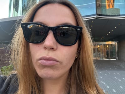 A woman wearing black sunglasses with the reflection of a building in the lenses faces the camera. She has fair skin and light brown hair. The background shows a modern building with glass windows and doors. The exterior includes a tiled surface with recessed lighting. The picture is taken at an unusual angle.