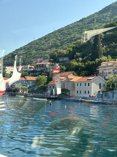 The image captures a waterside view of Lepetane, Montenegro, with a steep, tree-covered hillside rising behind a row of stone buildings. The foreground is dominated by the dark blue, rippling water of a bay, with several small white boats moored along a stone embankment. Some of these boats appear to be fishing boats, while others are smaller recreational vessels. A red and white buoy is visible in the water.
The buildings along the waterfront are constructed with stone and have red tile roofs. They vary in height and architectural style, with some appearing older and more traditional, while others are more modern. Several of these buildings have balconies and windows overlooking the water. Green foliage from trees and bushes is interspersed among the buildings and climbs up the hillside.
A few people can be seen on the embankment in front of the buildings, some appearing to be sitting or standing. A blue car is parked near one of the buildings. The sky is clear and blue, indicating a sunny day. The overall scene suggests a peaceful, picturesque coastal village.
