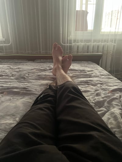 The media file captures a person's legs and feet resting on a bed, with a window and curtains in the background. The scene is set indoors, likely in a bedroom. The lighting suggests it's daytime, with sunlight streaming through the window and diffusing through sheer curtains, creating a soft, natural illumination. The bed appears unmade, with the duvet showing wrinkles and folds. The person is wearing dark-colored trousers. Their feet are bare, with one foot crossed over the other, indicating a relaxed posture. The window, with blinds and curtains, suggests a residential setting. There are no visible text elements in the image. The overall mood conveyed is one of relaxation and domestic comfort. The location context of Kaspiysk, Russia, while not explicitly visible, provides a geographical anchor to the scene.
