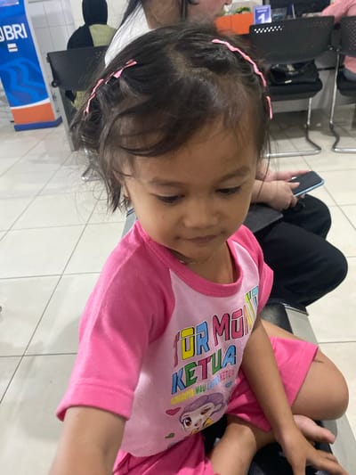 The image shows a young girl wearing a pink t-shirt with colorful text and a cartoon character. She has pink hair clips in her dark hair. The girl is lying down, and her face is turned away from the camera, with her eyes closed. It appears she may be sleeping or resting.

In the background, there are black chairs and what looks like a travel bag. Another person's hand is visible holding a mobile phone. The setting appears to be an indoor waiting area, possibly an airport or a train station, given the presence of seating and luggage. The floor is tiled.

The lighting suggests it's daytime, but there are no clear indicators of the weather. The overall mood is calm and relaxed, due to the child's posture.

On the t-shirt, there is text in multiple colors. The visible text includes "KETUA" which is Indonesian for "leader" or "chairman". There is also a cartoon illustration of a character with purple hair and pink eyes.

In the bottom left corner, part of a blue and orange sign is visible with the text "BRI", which is a major bank in Indonesia. This further supports the location being in Indonesia.