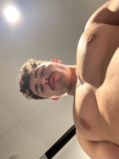 The image depicts an indoor scene in Chantepie, France, featuring one male individual viewed from a low-angle perspective. The subject, who appears shirtless, occupies the majority of the frame, with his head positioned towards the left and his muscular right shoulder and chest visible on the right. He has dark, curly hair, defined eyebrows, and a dark mustache. A thin, silver-colored chain necklace with a small, dark pendant rests on his upper chest. He is looking directly into the camera with a neutral expression. The background consists of a plain, light-colored ceiling with visible seams or panels. A bright, artificial light source is visible in the upper left corner, and a dark, linear structural element is present in the lower right background.