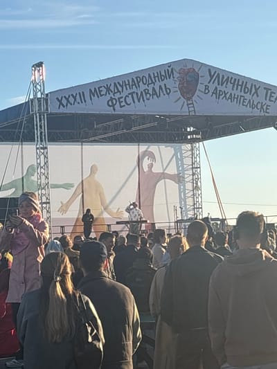 This image captures an outdoor event, likely a festival, with a large banner visible on a stage structure. The banner displays text in Russian and artwork. The text on the banner reads "УЛИЧНЫХ ТЕ" and "В АРХАНГЕЛЬСКЕ" on the top portion, and "РОДНЫЙ МЕЖДУНАРОДНЫЙ ФЕСТИВАЛЬ" with "XXXII" preceding it. The artwork includes a stylized, red, clenched fist with a ladder ascending from it, and at the bottom, illustrations of human figures with outstretched arms.
A crowd of people are gathered, seemingly watching or participating in the event. They are mostly seen from behind or in profile, with their faces largely obscured. The attire of the attendees suggests a casual, outdoor setting. The lighting indicates it is daytime, with bright sunlight casting shadows and highlighting the scene. The sky is clear and blue, suggesting fair weather. The overall impression is of a public gathering, possibly a cultural or artistic festival, taking place in Arkhangelsk, Russia, as indicated by the text on the banner. The presence of a stage and banner implies a performance or a formal presentation.