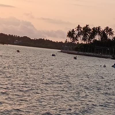 The image depicts a body of water at sunset, likely a lake or bay, with several people engaging in recreational activities. In the distance, there are trees lining the horizon and a few buildings visible. Closer to the right side of the image, there's a row of palm trees along the shoreline, and a concrete barrier or walkway.
There are at least three kayaks or canoes on the water, each carrying one or two people, and some appear to be paddling. Further to the right, two individuals are swimming.
The scene appears tranquil, with a focus on leisure activities during the sunset hours. The location is identified as Badem, India; this is indicated for location purposes only and is not visually verifiable from the image itself.