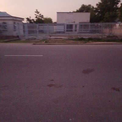 The image shows a street scene in Wuro Allahamdu, Nigeria. In the background, a low-rise building with a light-colored facade is visible, partially obscured by a metal fence. Behind the fence, more buildings and trees can be seen. The fence itself appears to be made of vertical metal bars. The foreground is dominated by an asphalt road with a single white line marking the lane. There are a few dark, irregular stains on the road surface. The sky is overcast and a pale, dusky color, suggesting it is either dawn or dusk. The overall impression is of a quiet, rural or semi-urban setting. There are no people or vehicles clearly visible in the frame. The lighting is dim, indicating low natural light conditions. No text is discernible in the image.