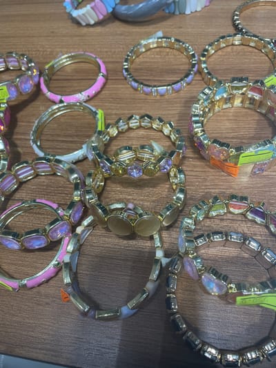 The image shows a close-up, overhead view of several gold-toned bracelets arranged on a wooden surface. The bracelets are diverse in design, featuring various colorful gemstones, beads, and enamel accents in shades of pink, purple, blue, and white. Some bracelets have a delicate chain-like construction, while others are made of larger, faceted stones. Price tags are visible on a few of the bracelets, with one clearly showing "550.00". The background is consistent with a display in a retail environment. The image does not provide any information about the specific location within Yunus Emre, Turkey.