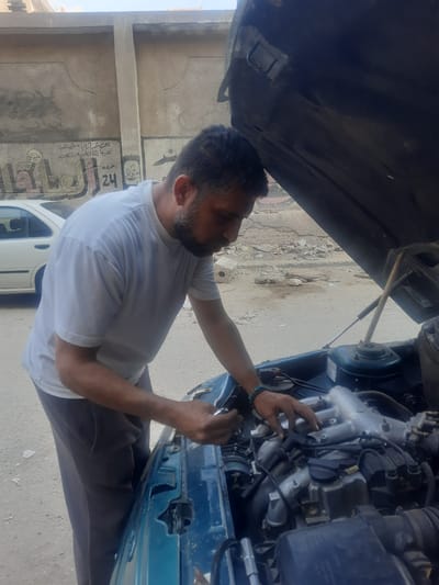 A man with a beard is working on a car engine. The car hood is open, and the man is reaching into the engine compartment. The car appears to be parked on a street in front of a building with faded Arabic script painted on the wall. The location is likely in Cairo, Egypt based on the architectural style of the buildings and the written script.