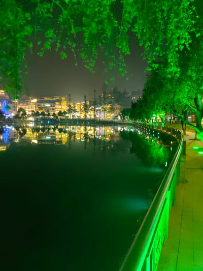 The image depicts a nighttime scene at Sa Pa Lake in Sa Pa, Vietnam. The foreground is dominated by a dark body of water, Sa Pa Lake, which occupies the central and left portions of the frame. Overhanging foliage, illuminated with a vibrant green light, partially obscures the top of the image. Along the right side, a paved pedestrian walkway curves around the lake, lit by multiple circular green in-ground lights. A metal handrail, also bathed in green light, runs parallel to the walkway, separating it from the water.
Across the lake, a cluster of multi-story buildings forms the background, brightly illuminated with warm yellow and white lights, which reflect distinctly on the dark lake surface. One building on the far bank is identifiable by the text "SAPA PARADISE HOTEL" visible on its facade. Numerous trees, appearing both deciduous and coniferous, line the far bank and are interspersed among the buildings. On the far left bank, a prominent, large structure illuminated with blue lights stands, its reflection also visible in the lake. The sky above the buildings is dark and overcast. No individuals or specific actions are discernible within the frame.