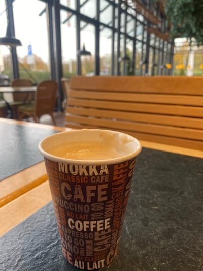 A disposable coffee cup containing a light brown beverage with a frothy top is positioned in the foreground, slightly right of center, on a dark, textured tabletop. The cup's exterior features a repeating pattern of white text on a brown and orange background, including words like "MOKKA," "CLASSIC CAFE," "CAFE," "COFFEE," "CAPPUCCINO," and "ESPRESSO." To the left of the cup, a section of a lighter brown wooden surface is visible on the table.

The midground features interior elements of what appears to be a cafe or restaurant. A long, light-brown wooden bench with horizontal slats runs across the scene, positioned behind the table. Further left, another table with two rattan-style chairs is visible, partially obscured.

The background consists of large, multi-paned glass windows with dark frames, extending vertically and horizontally, providing a view of the outdoors. Outside, blurry trees with hints of yellow and orange foliage suggest autumn conditions. A blurred blue and white street sign and a vehicle are faintly discernible through the left window pane. Inside, dark, bell-shaped pendant lights hang from the ceiling. Decorative green foliage, possibly artificial, hangs from a beam or ceiling structure on the right side of the background. No people are visible, and no actions or interactions are depicted. The environment is indoor, well-lit by natural light through the windows, suggesting a daytime scene. The specific location within Lichtenau, Germany, cannot be determined from the visual information provided in the image.