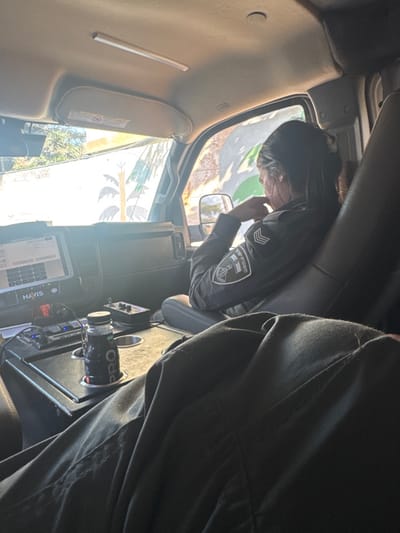 The image depicts the interior of a vehicle, viewed from behind the front seats, focusing on the passenger side and the exterior environment. A female individual, identified as an Israel Police officer by the "משטרת ישראל" (Israel Police) patch and chevrons on her dark uniform sleeve, is seated in the front passenger seat. Her dark hair is pulled back, and her right hand is positioned near her chin, suggesting contemplation or repose. The legs of another individual, wearing dark pants, are visible in the foreground, indicating a person is seated in an adjacent position, likely the driver's seat.

The vehicle's dashboard features a digital display with "HAVIS" visible on its lower left. A center console between the front seats contains cup holders, one of which holds a dark bottle labeled "PRO." Additional electronic controls and devices are present on the console.

Through the windshield and passenger window, bright daylight illuminates a light-colored exterior wall adorned with a mural depicting stylized green and dark-colored palm tree-like shapes. Green foliage is visible beyond this wall. This scene is located in the city of Lod, Israel.