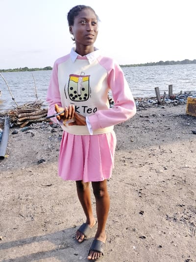 A young woman stands near a shoreline, dressed in a light pink pleated skirt and a cream-colored sweatshirt with pink sleeves. The sweatshirt features a "Milk Tea" graphic. She wears black sandals and holds a dark-colored object in her hands. The background reveals a body of water, driftwood, and scattered debris. The scene is captured in daylight.