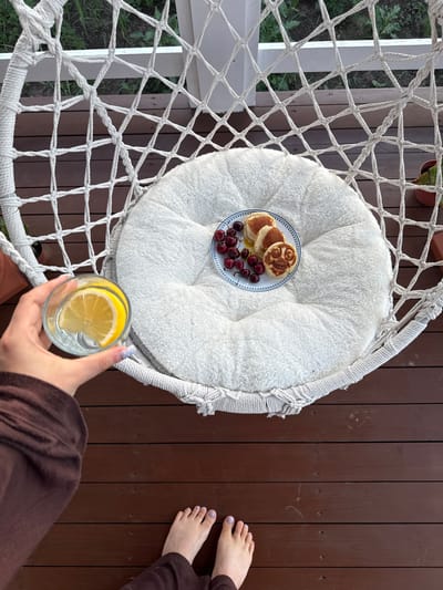 An overhead shot captures a person's hand holding a glass of water with a lemon slice, positioned on the right side of a hammock chair on a wooden deck. Inside the chair, on a white circular cushion, sits a plate of pancakes and cherries. The pancakes on the plate are arranged to resemble faces, with syrup drizzled over them. The surrounding environment includes a white railing and greenery in the background. The scene appears to be a relaxed outdoor setting, possibly on a porch or balcony in Starokorsunskaya, Russia, given the context.