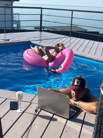 A person with dark curly hair, wearing a baseball cap and a white bracelet, is sitting by a swimming pool and looking at a laptop. The person is on a wooden deck, with a pool filled with blue water to their left. The person's right arm is resting on the laptop, and their left hand is near their face. A pink inflatable flamingo float is in the pool, near another person whose legs and torso are visible. This person is in the water, holding onto the flamingo, and appears to be sunbathing or relaxing. The scene is outdoors, suggesting a warm, sunny day, likely daytime given the bright lighting and clear blue sky reflected in the water. The background shows the calm, blue expanse of the sea or a large body of water. In the foreground on the deck, there is a white champagne flute and a dark object, possibly a phone or a small remote, placed next to the laptop. The wooden deck is made of grey planks. The overall atmosphere is one of leisure and relaxation, with a touch of work potentially being done despite the vacation-like setting. There is a visible logo on the laptop that reads "Lenovo". The cap worn by the person has a patch with an eagle or bird of prey.